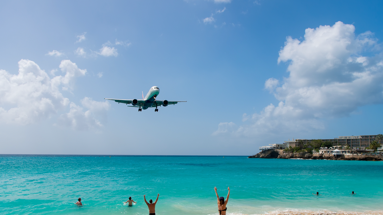 Avion qui vole au-dessus d'une plage des Caraïbes, avec des personnes dans l'eau qui lèvent les bras en l'air.
