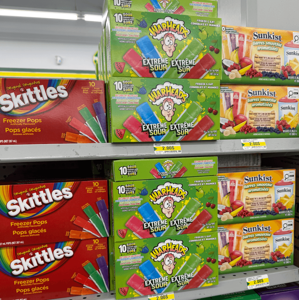Bo\u00eetes de pops glac\u00e9s Skittles et Warheads expos\u00e9es sur une tablette de Dollarama.