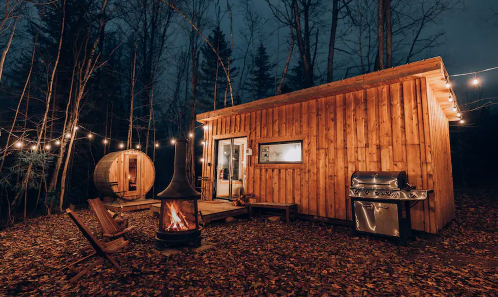 Cabane en bois entour\u00e9e d'arbres et d'une guirlande lumineuse, avec un foyer, des chaises, un barbecue et un sauna sur le terrain.