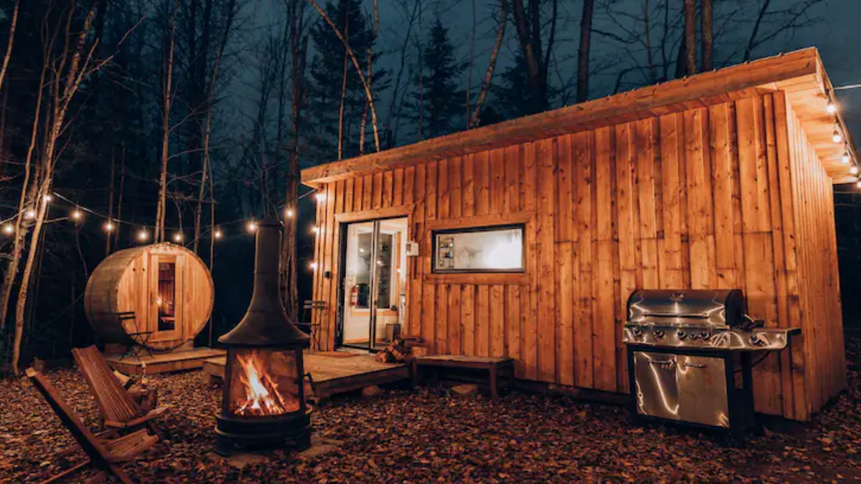 Cabane en bois entourée d'arbres et d'une guirlande lumineuse, avec un foyer, des chaises, un barbecue et un sauna sur le terrain.