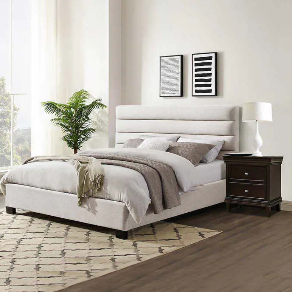 chambre \u00e0 coucher avec tr\u00e8s grand lit en tissu beige, table de chevet brune, plante et tapis.