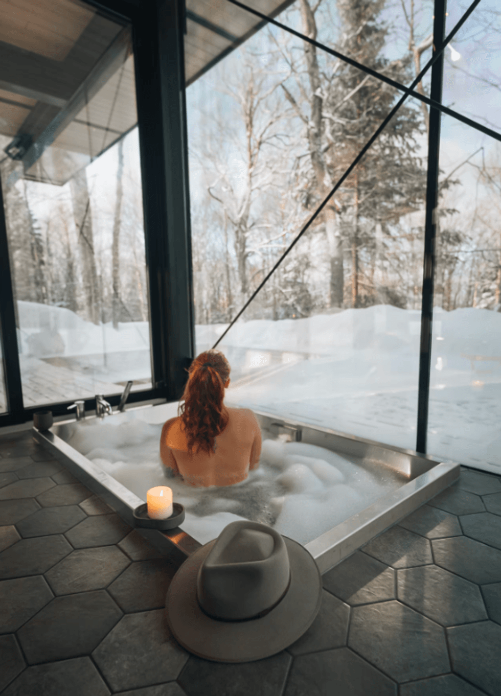 Femme dans un bain moussant encastr\u00e9 dans le plancher, \u00e0 c\u00f4t\u00e9 d'une chandelle et d'un chapeau, face \u00e0 de grandes vitres donnant sur la for\u00eat enneig\u00e9e.