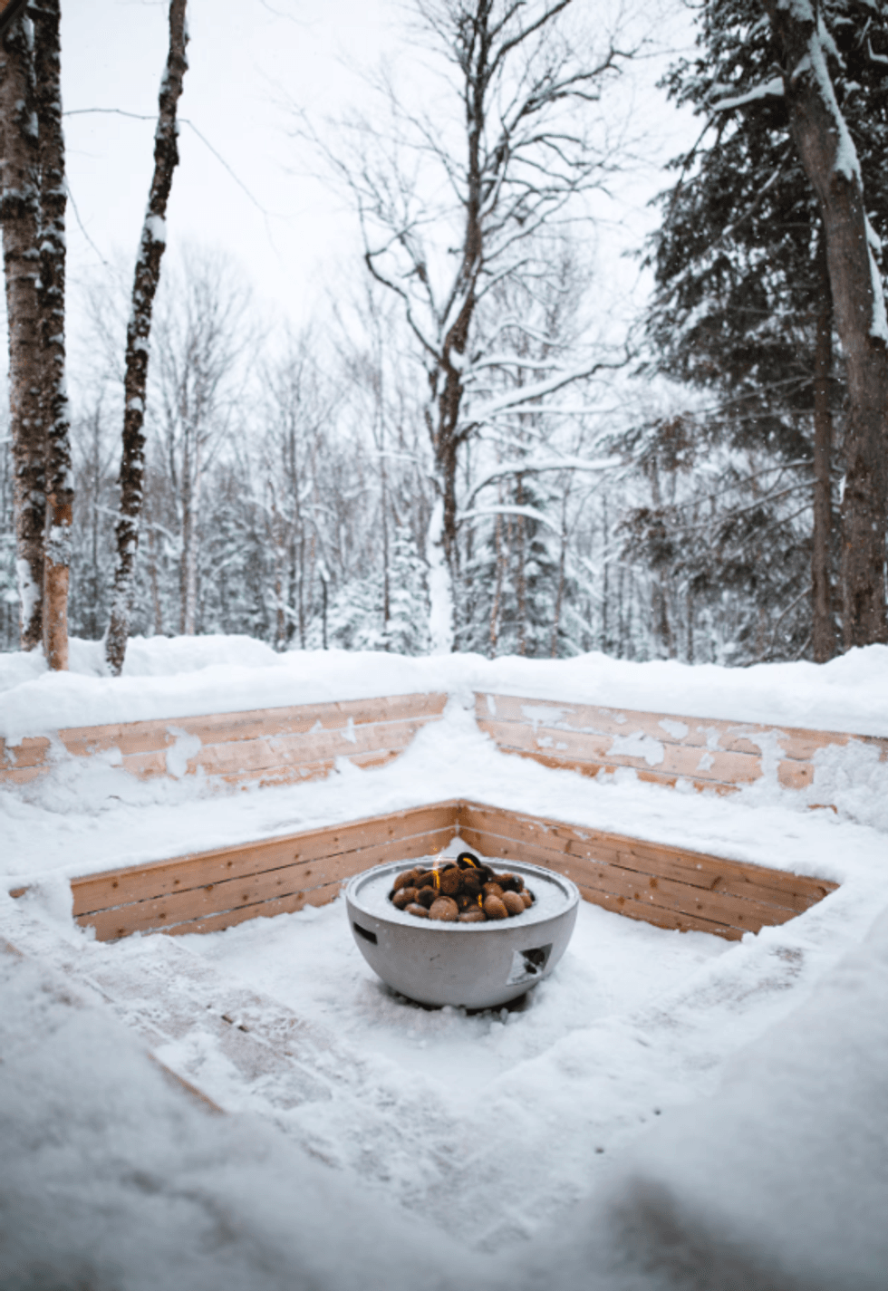 Foyer ext\u00e9rieur rond en b\u00e9ton au centre d'un coin am\u00e9nag\u00e9 avec des bancs de bois recouverts de neige, en pleine for\u00eat.