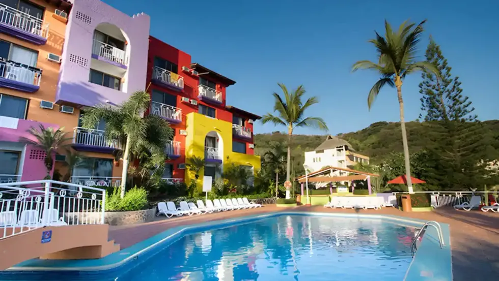 Piscine ext\u00e9rieure entour\u00e9e de palmiers et de b\u00e2timents color\u00e9s, avec vue sur les collines verdoyantes de la Riviera Nayarit, au Decameron Los Cocos Guayabitos Ramada.