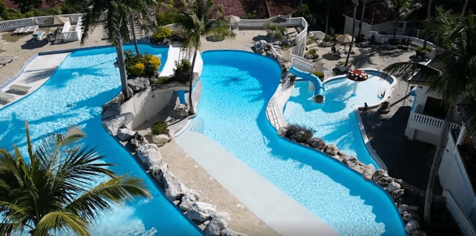 Piscines ext\u00e9rieures sinueuses avec zones peu profondes, palmiers et glissades d\u2019eau dans un complexe tout inclus.