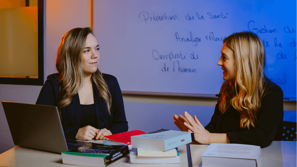 \u00c9tudiante discutant avec son enseignante autour d\u2019une table avec des livres et un ordinateur portable dans une salle de travail.