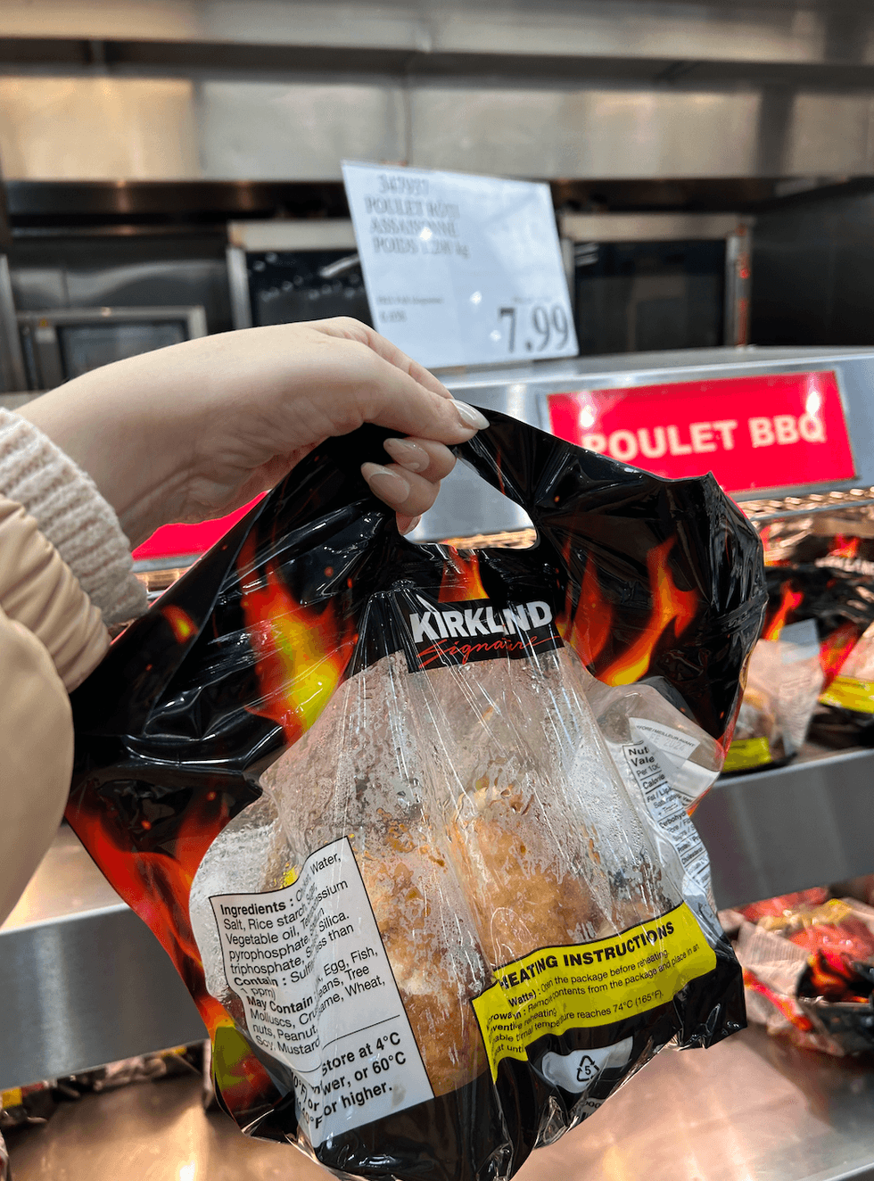 Une main tenant un sac de poulet r\u00f4ti Kirklanf Signature devant le pr\u00e9sentoir chauff\u00e9 du Costco.