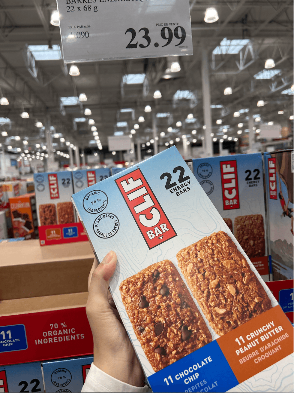 Une main tenant une boite de barres \u00e9nerg\u00e9tiques Clif Bar au p\u00e9pites de chocolat et au beurre d'arachide croquant devant un \u00e9talage du Costco.
