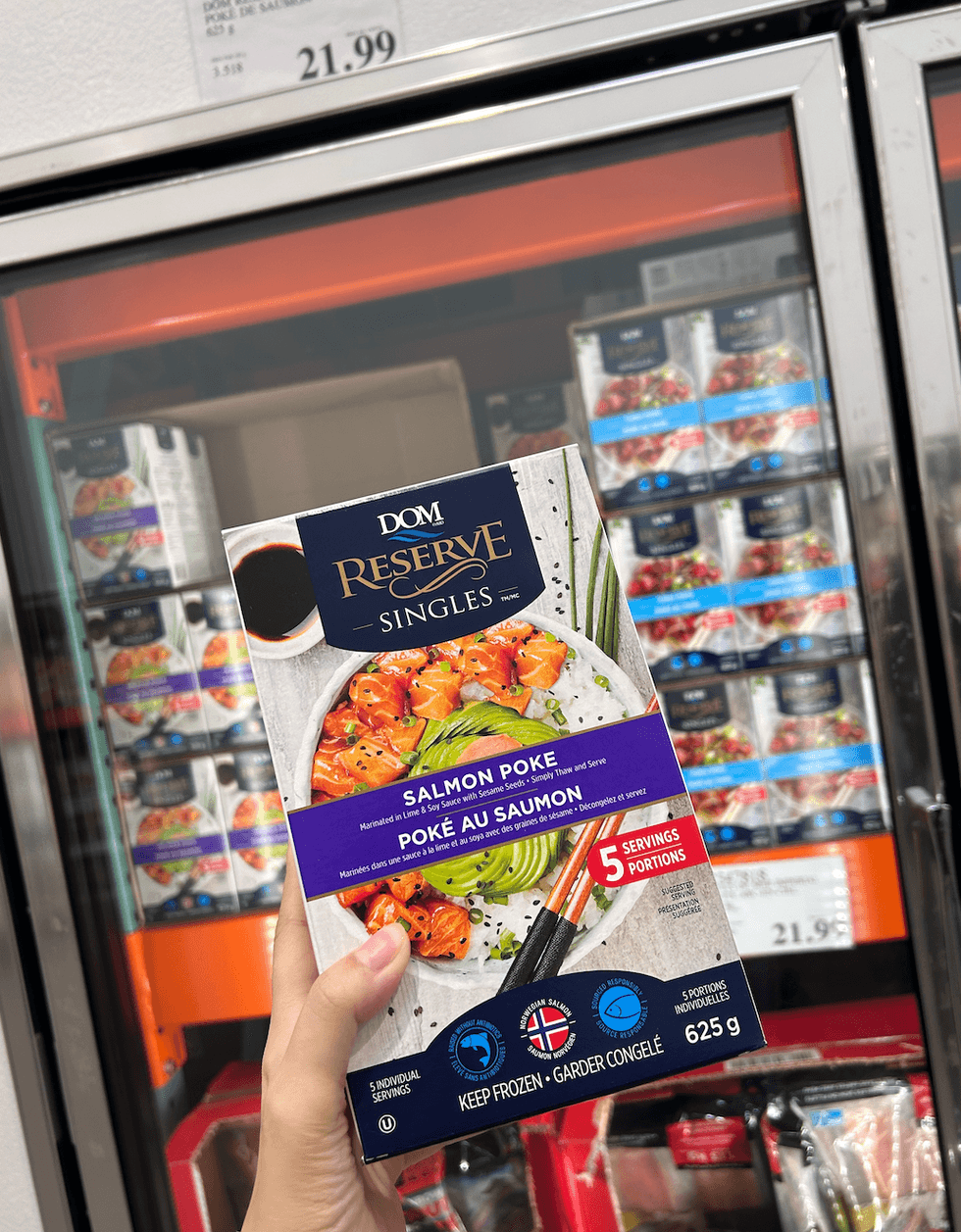 Une main tenant une boite de pok\u00e9 au saumon DOM Reserve devant un \u00e9talage du Costco.