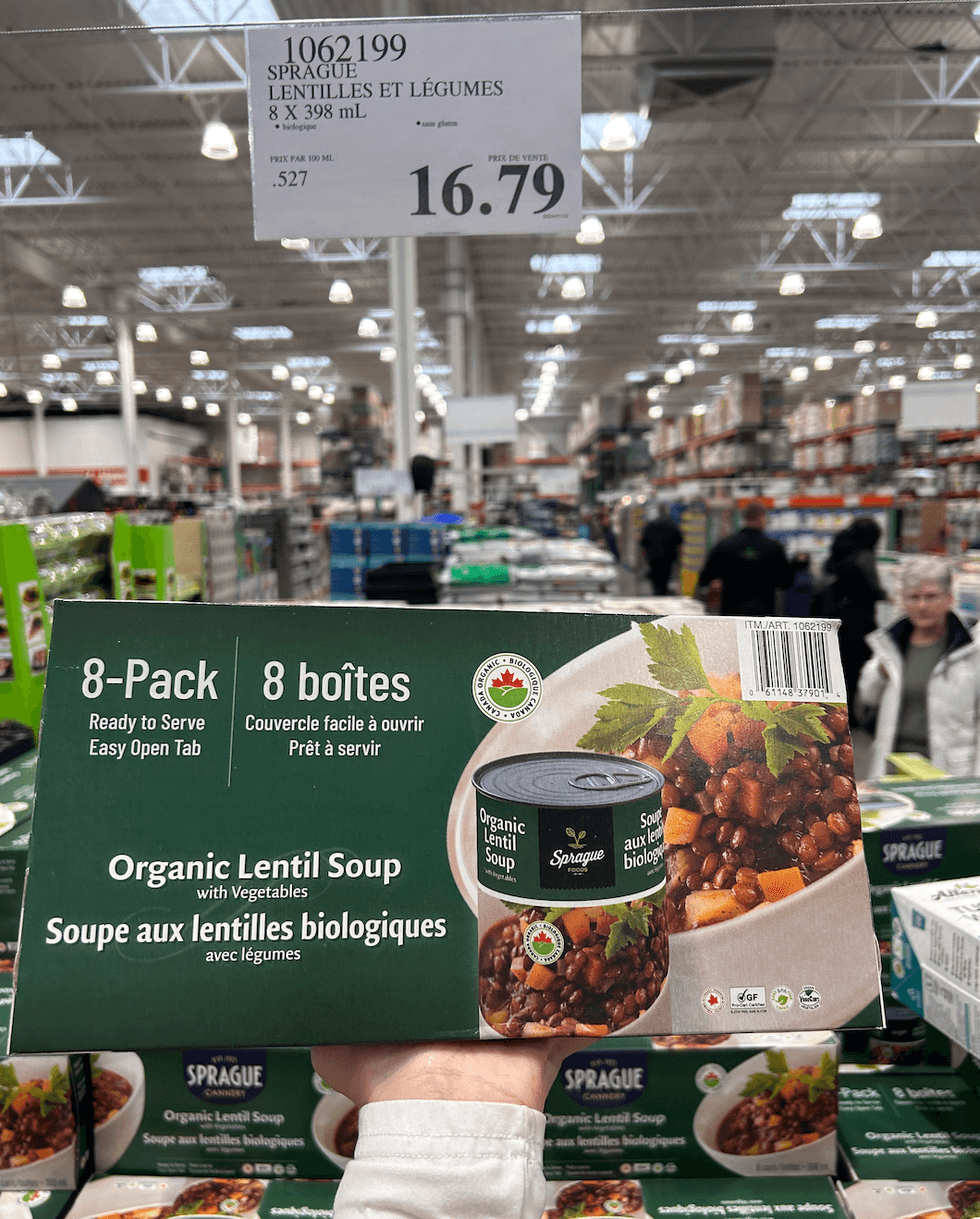 Une main tenant une boite des soupes aux lentilles et l\u00e9gumes de la marque Sprague devant un \u00e9talage du Costco.