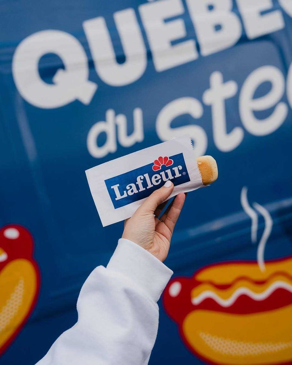 Une main tient un steam\u00e9 dans un emballage Lafleur devant une affiche o\u00f9 l\u2019on peut lire \u00ab Journ\u00e9e qu\u00e9b\u00e9coise du steam\u00e9 \u00bb.