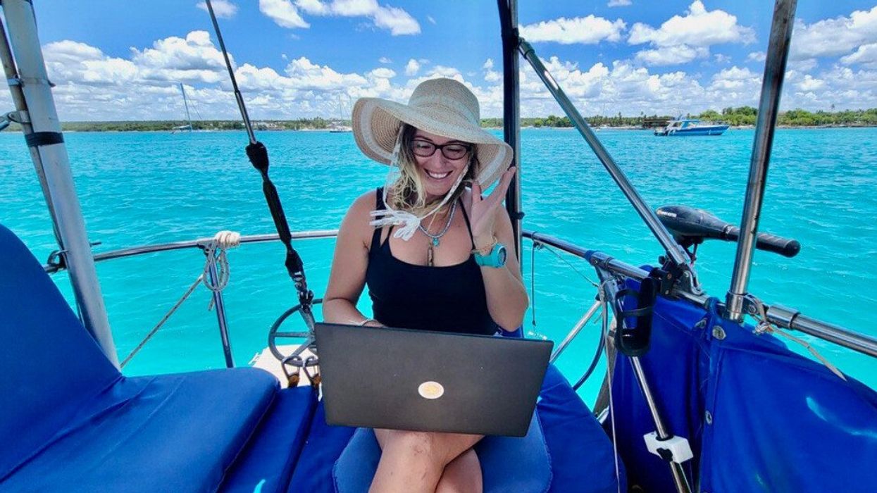 Une personne est assise avec son ordinateur portable sur les genoux à bord de son voilier avec vue sur la mer en arrière-plan.