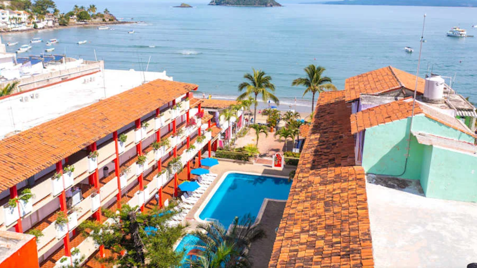 Vue a\u00e9rienne du Decameron Los Cocos Guayabitos Ramada, avec sa piscine centrale, ses b\u00e2timents color\u00e9s et un acc\u00e8s direct \u00e0 la plage de Guayabitos, face \u00e0 la baie.