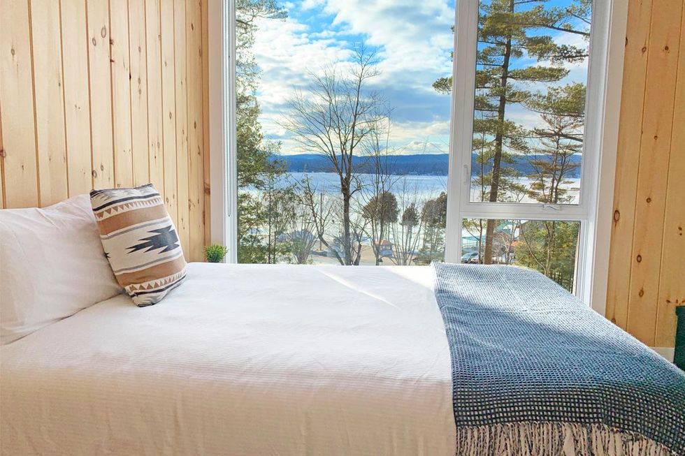 Vue de c\u00f4t\u00e9 d'un lit dans le chalet, avec vue des arbres et du lac en arri\u00e8re-plan.