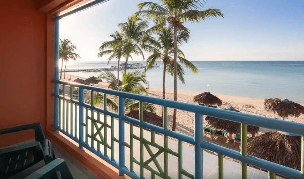 Vue depuis un balcon d\u2019h\u00f4tel aux murs orang\u00e9s et \u00e0 la rambarde turquoise et verte, donnant directement sur une plage.